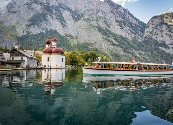 Boot auf dem Königssee mit kleinem Ort und Bergen im Hintergrund.